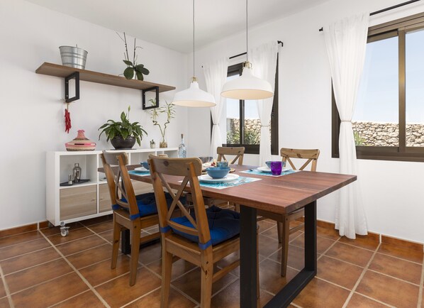 A dining area with a wooden table, four chairs, and a white pendant light. There are shelves above the table holding plants and decorative items.