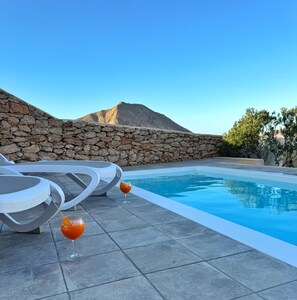 A pool area with stone walls, lounge chairs, and two glasses of orange drink.