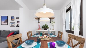 Un coin repas avec une table en bois dressée pour six personnes, entourée de chaises. Des tableaux encadrés ornent le mur, un luminaire suspendu éclaire la pièce, et une fenêtre est agrémentée de rideaux.