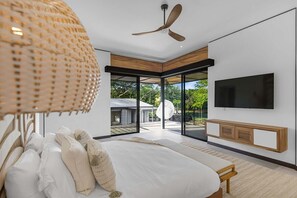 A modern bedroom with a large bed, a ceiling fan, a TV mounted on the wall, and a view of the outdoors through a glass door.