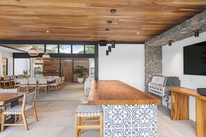 An open-plan dining area with a long wooden table and chairs, a stone accent wall, and a built-in grill station.