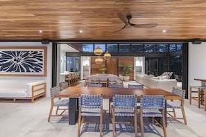 A modern dining area with a wooden table and chairs, a ceiling fan, and a large window with a view of the outdoors. - Casa Que Chiva (Reserva de Golf, Hacienda Pinilla)