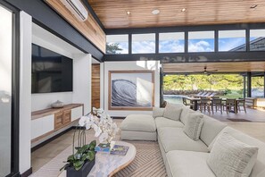 Ein modernes Wohnzimmer mit einer großen Couch, einem Flachbildfernseher und Blick auf einen Pool und Grünflächen.