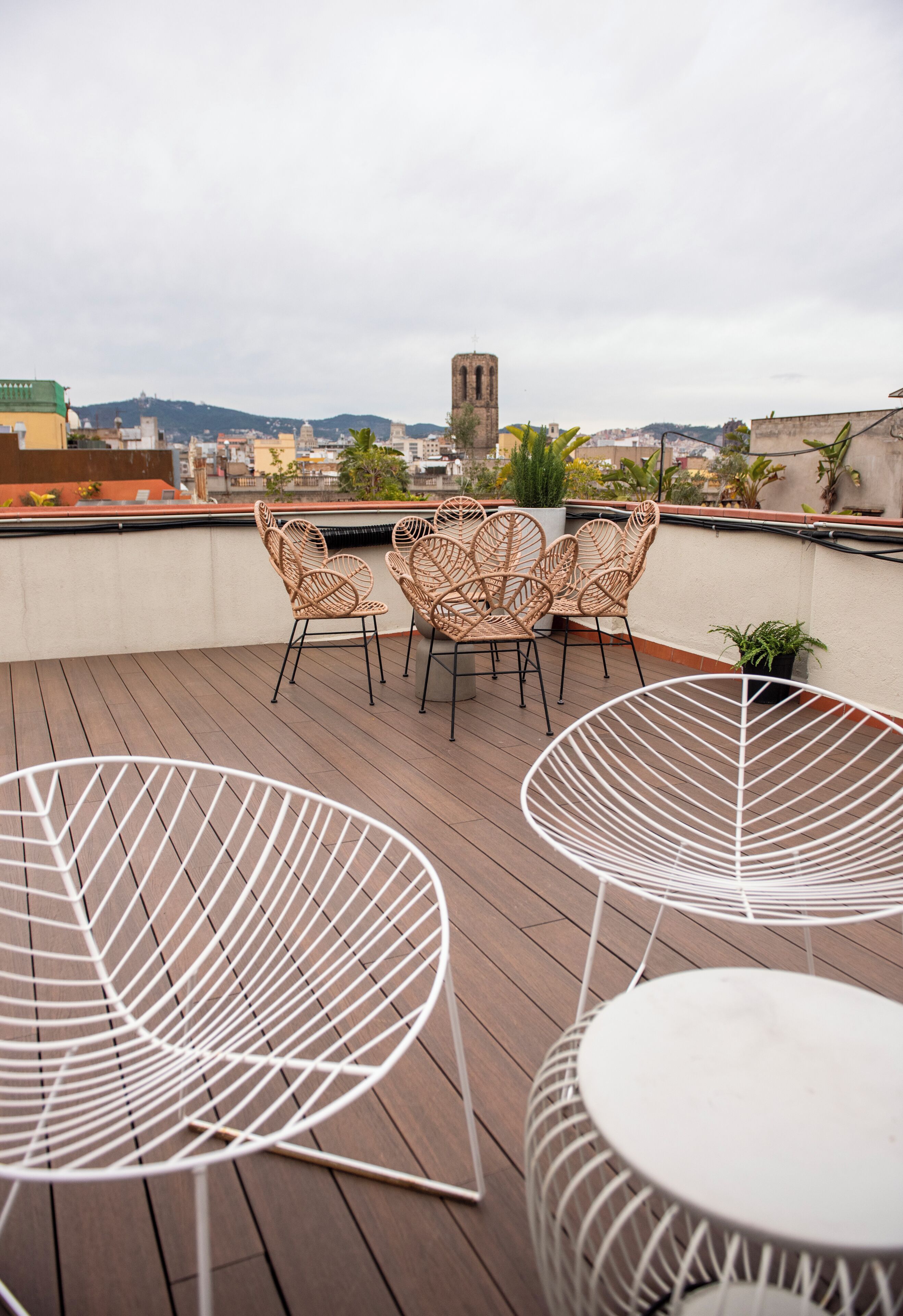 Une terrasse sur le toit, avec des fauteuils en osier et des chaises en fil de fer blanc, une vue sur le paysage urbain avec le clocher d’une église, et une terrasse en bois.