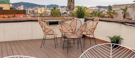 A rooftop terrace with wicker chairs and white wire chairs, a view of a cityscape with a church tower, and a wooden deck.