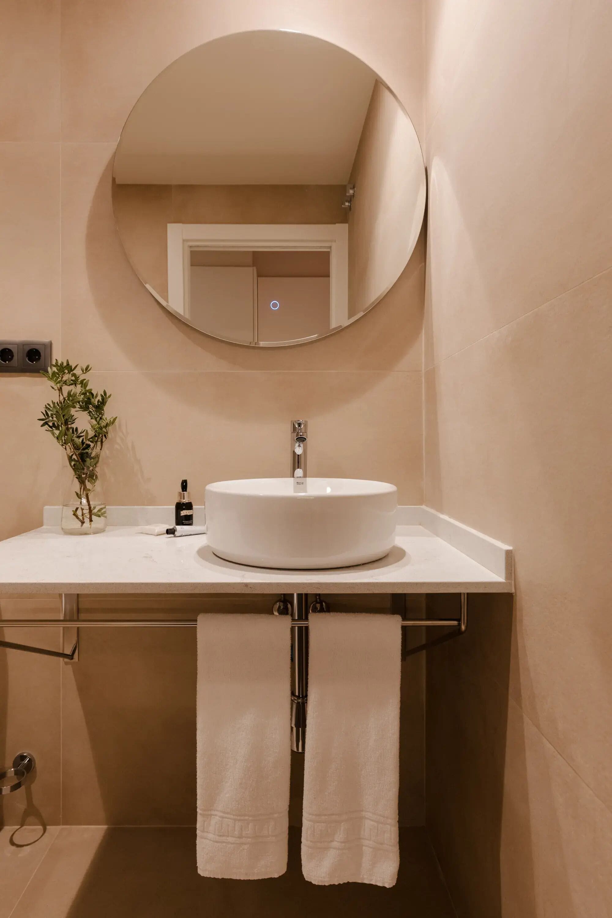 Une salle de bain avec un miroir rond, un lavabo blanc, un vase avec des plantes vertes et deux serviettes suspendues.
