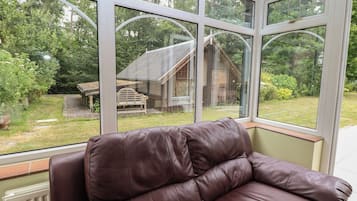 Quarto com sofá de couro marrom posicionado perto de grandes janelas com vista para um jardim com estrutura de madeira e um banco.