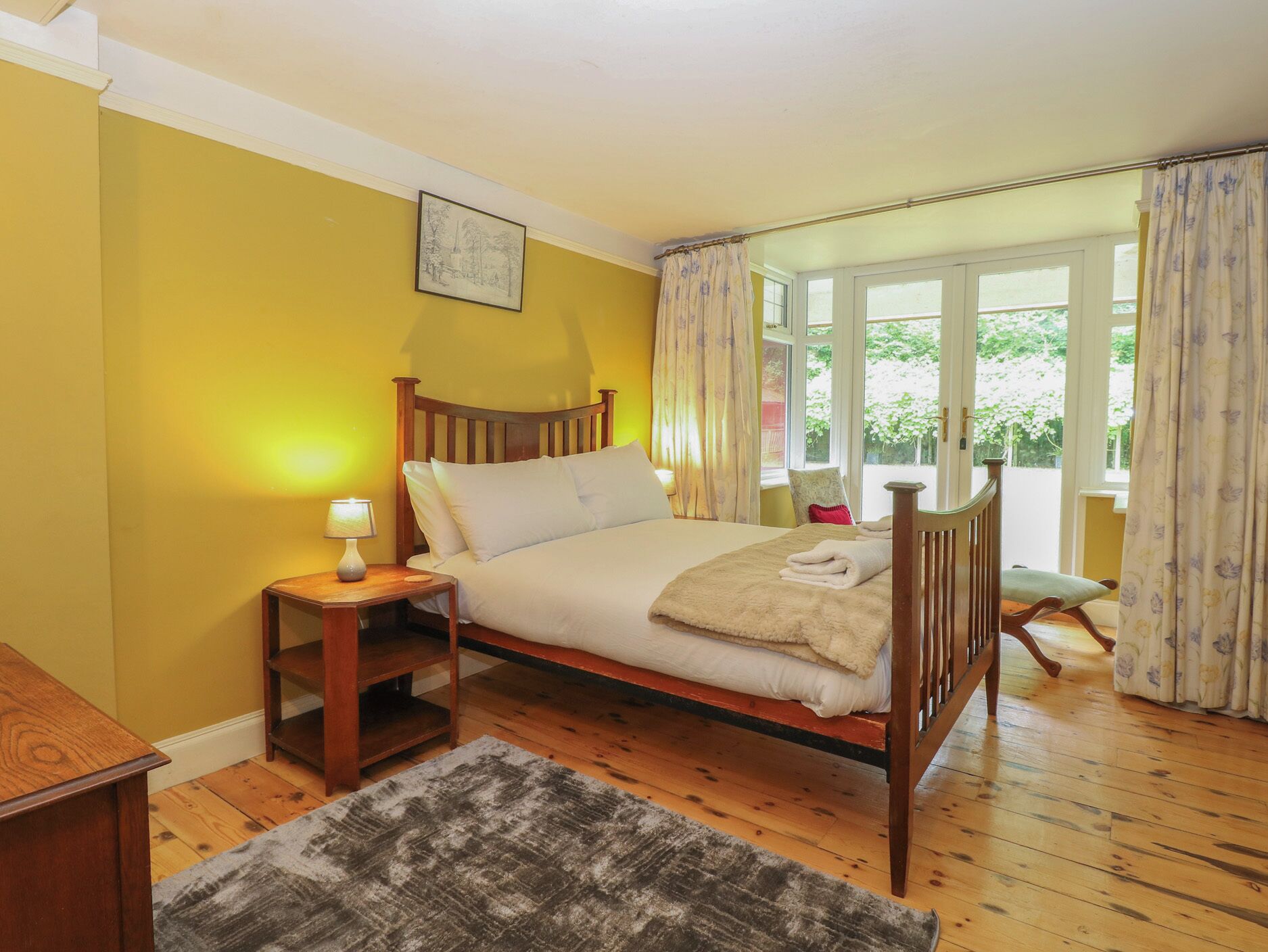 A bedroom with a wooden bed, a bedside table with a lamp, a window with curtains, and a view of greenery outside.
