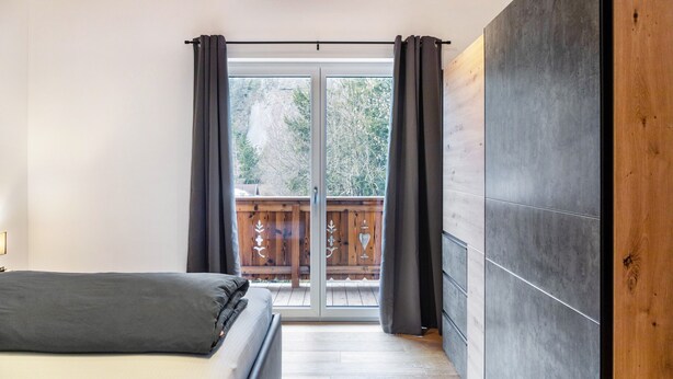 A bedroom with a bed, a sliding glass door to a balcony, and a wooden ceiling.