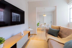 Ein modernes Wohnzimmer mit einem an der Wand befestigten Flachbildfernseher, einem Holztischelement, einem beigefarbenen Sofa mit Kissen und einem Essbereich im Hintergrund.