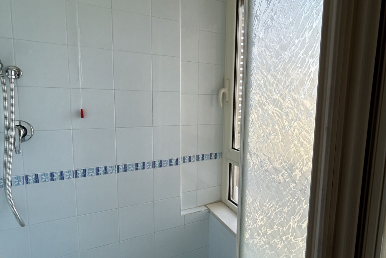Une salle de bain avec des murs recouverts de carreaux blancs, un espace douche et une fenêtre donnant sur l’extérieur.