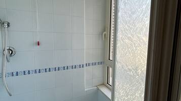 Une salle de bain avec des murs recouverts de carreaux blancs, un espace douche et une fenêtre donnant sur l’extérieur.