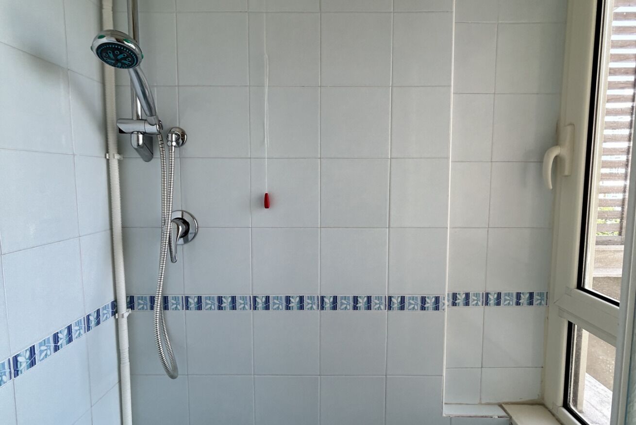 Een badkamer met witte betegelde wanden, een douche met handdouche en een raam met blinds.