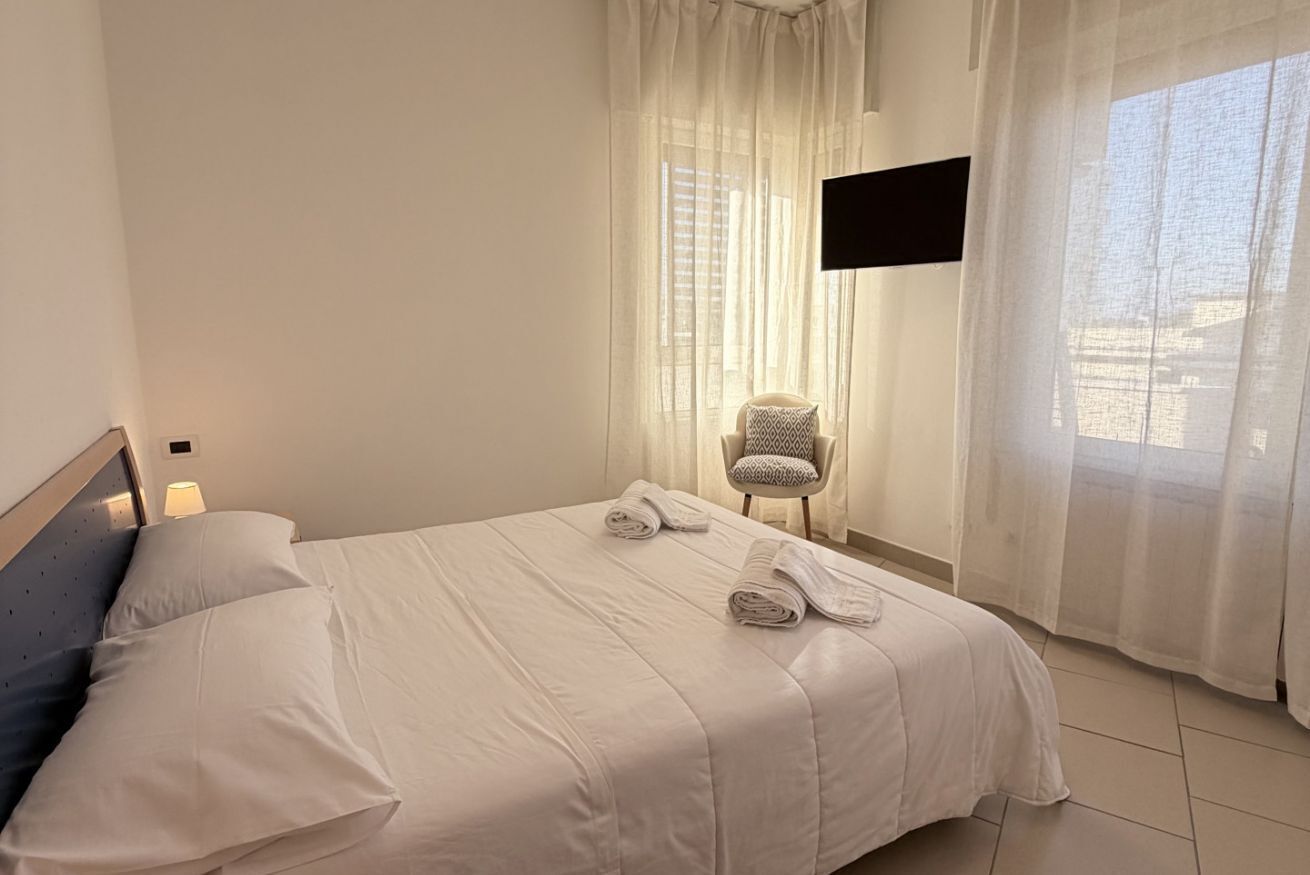 Un lit bien fait, avec du linge de lit et des serviettes blanches, une chaise agrémentée d’un coussin à motifs et un téléviseur fixé au mur.