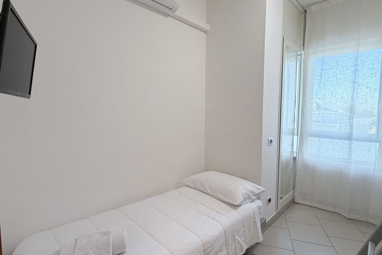 Quarto pequeno com uma cama de solteiro, edredom, travesseiro e toalha brancos. Há uma televisão de tela plana montada na parede à esquerda da cama. O quarto possui uma porta de vidro deslizante que dá para uma varanda com vista da cidade.