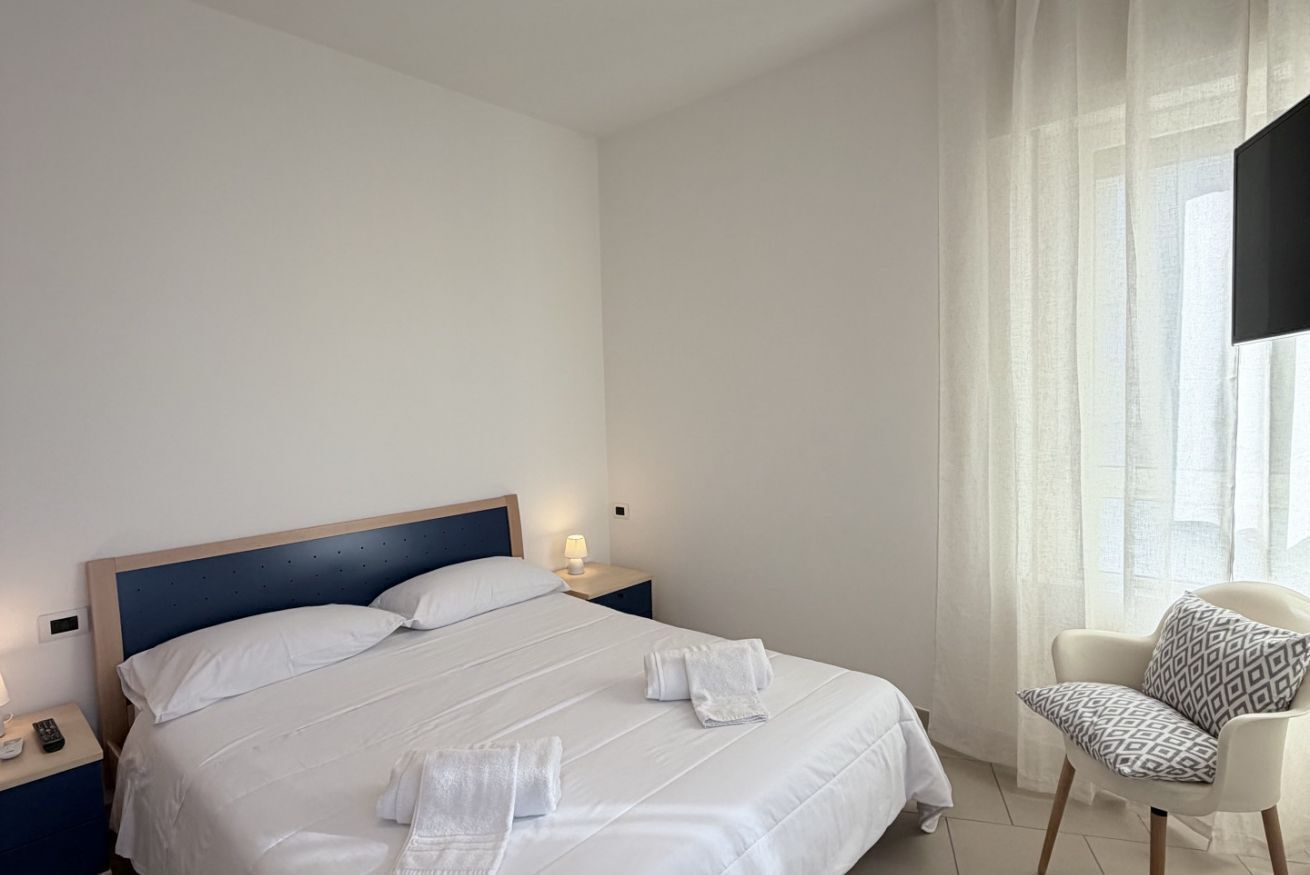Un lit bien fait, avec du linge de lit et des serviettes blanches, une table de chevet avec une lampe et une chaise agrémentée d’un coussin à motifs.