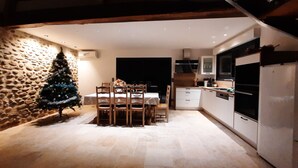 A modern kitchen with white cabinets, a built-in oven, and a dining area with a table and chairs. A small Christmas tree is visible in the corner.