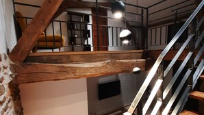 A spacious loft-style room with exposed wooden beams, a mezzanine level, and a staircase with a metal railing.