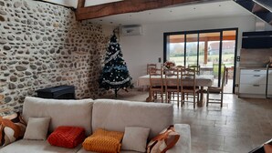 A cozy living room with a stone wall, a decorated Christmas tree, a sofa with cushions, a dining area with a table and chairs, and a kitchen with cabinets.