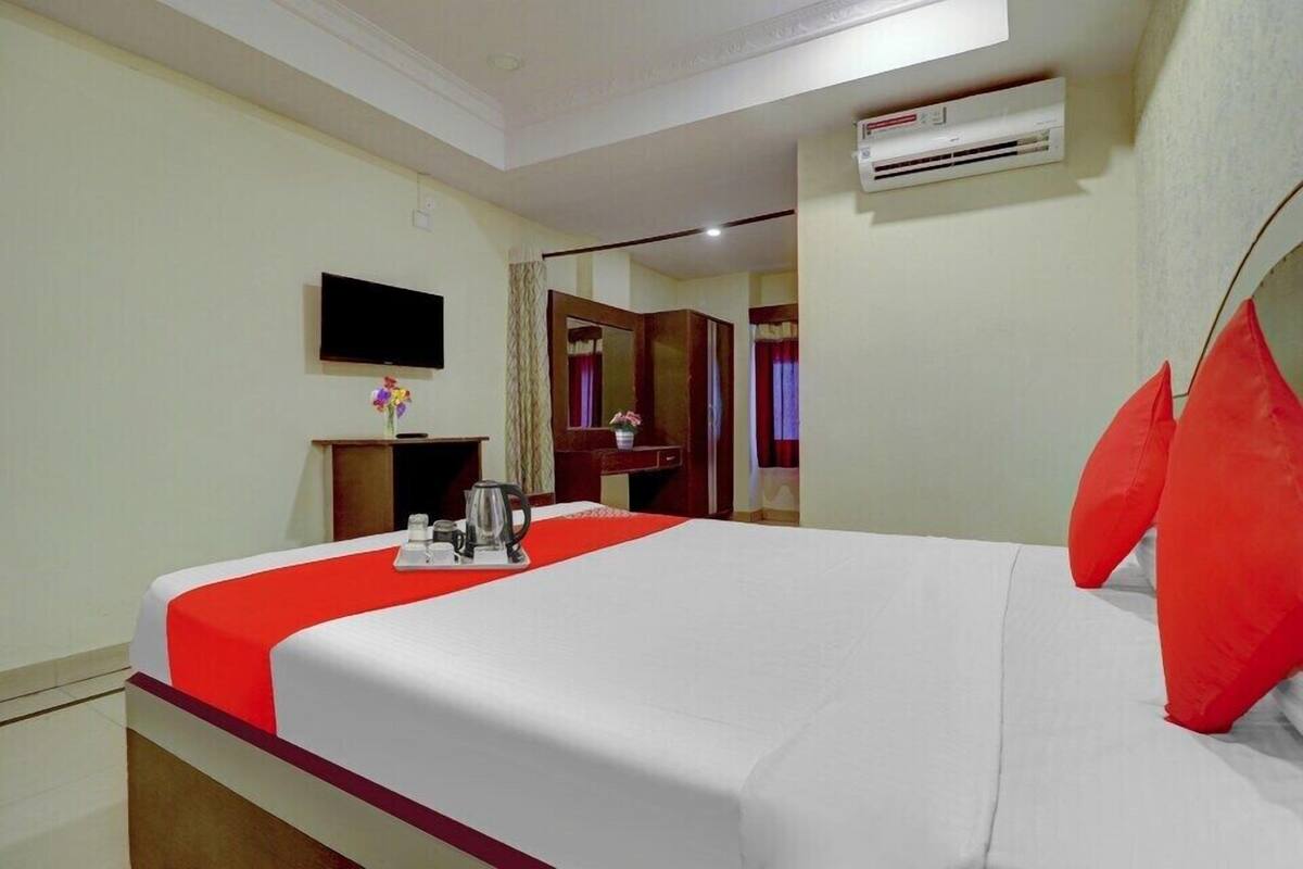 A hotel room with a bed, a television, an air conditioning unit, and a small table with a kettle and cups.