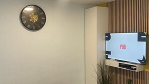 A modern living room with a wall-mounted clock, a flat-screen TV, a small white table, and a couple of potted plants. - Bagnol Cosy - Cosy apartment for 3 people, 30 min from Avignon (Bagnols-sur-Cèze)