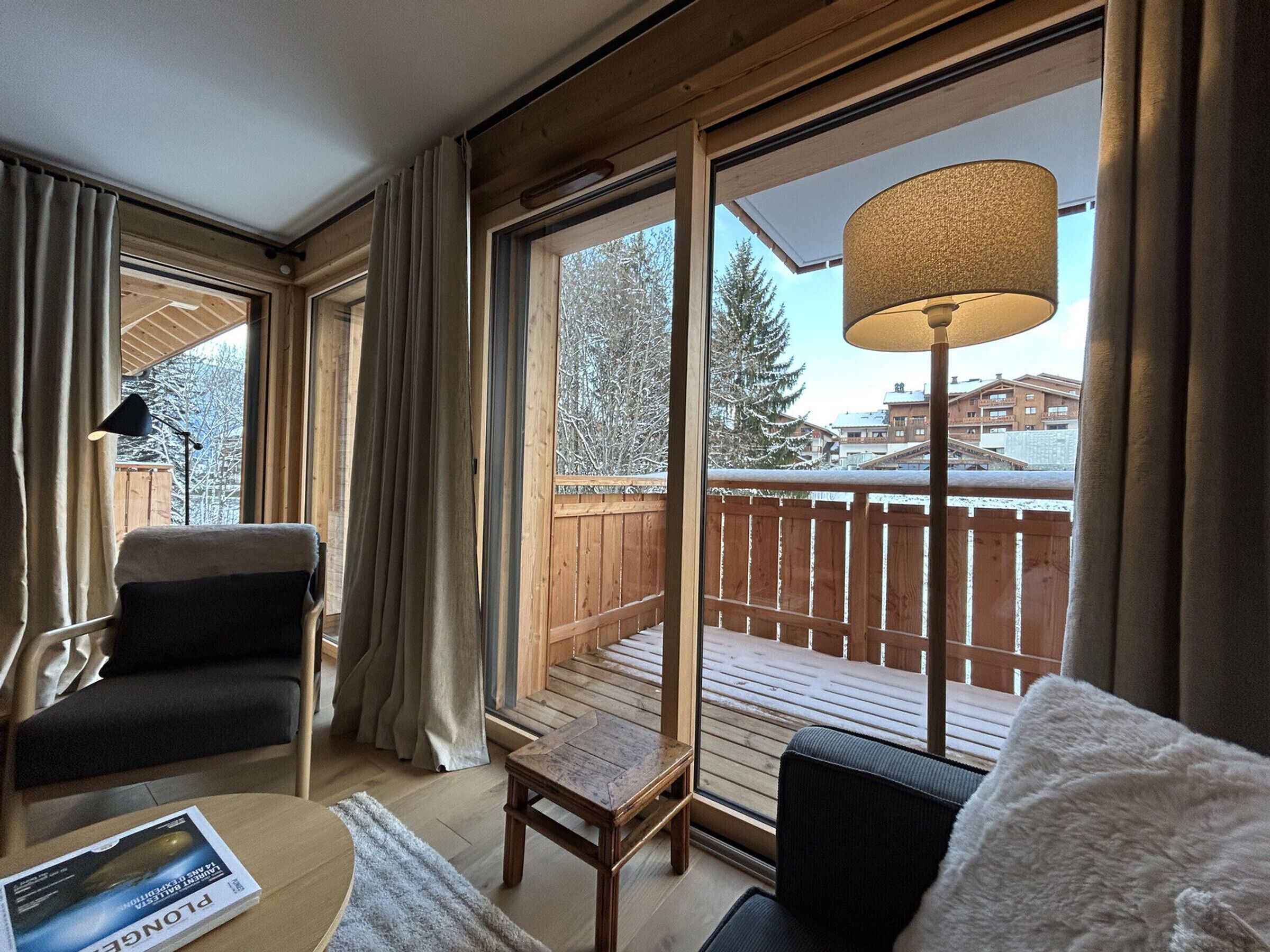 Ein gemütliches Zimmer mit einer Holzterrasse, einem großen Fenster mit Vorhängen, einer Lampe und einem Stuhl.