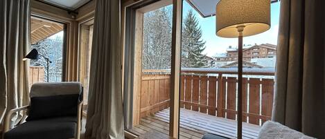 Ein gemütliches Zimmer mit einer Holzterrasse, einem großen Fenster mit Vorhängen, einer Lampe und einem Stuhl.