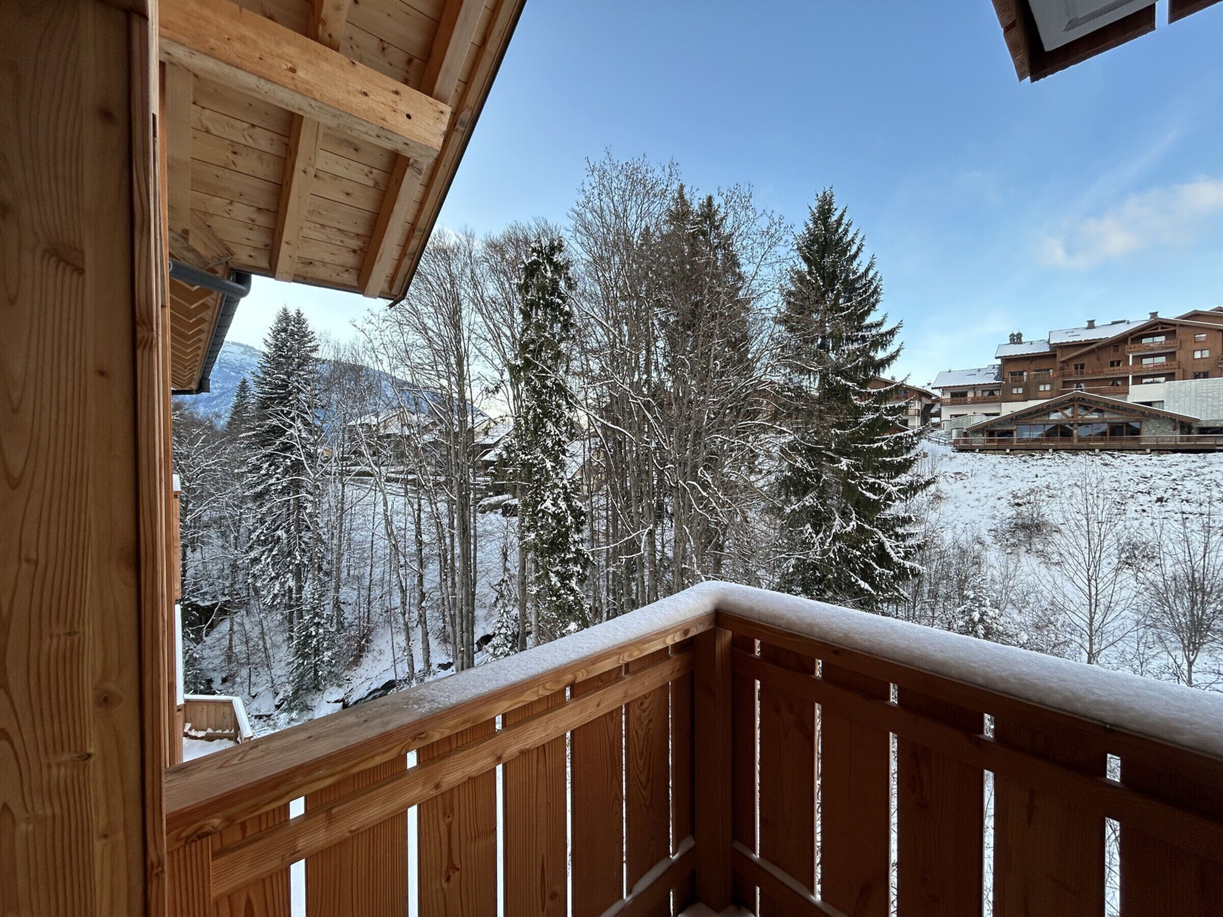 A wooden balcony with a view of snow-covered trees and buildings in the background.