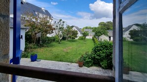 Vista a través de una ventana que muestra un patio con césped, plantas en macetas y un edificio con techo inclinado.