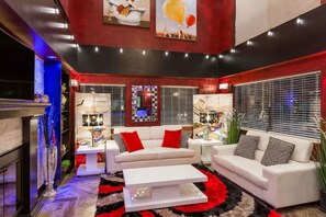 Uma sala de estar moderna com sofás brancos, almofadas vermelhas e um tapete preto e vermelho. Há duas mesas laterais com luminárias, uma lareira e uma janela grande com persianas.