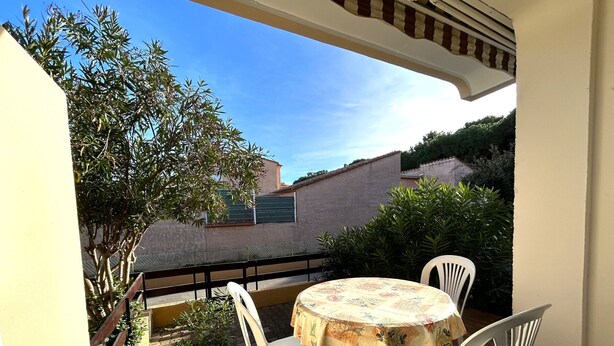 Une terrasse avec une table ronde et quatre chaises, donnant sur un jardin et un bâtiment entouré d’une clôture verte.