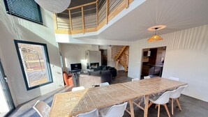 Un vaste espace de vie comprenant une longue table en bois et des chaises blanches, un escalier menant à un niveau supérieur, et une grande fenêtre laissant entrer la lumière naturelle.