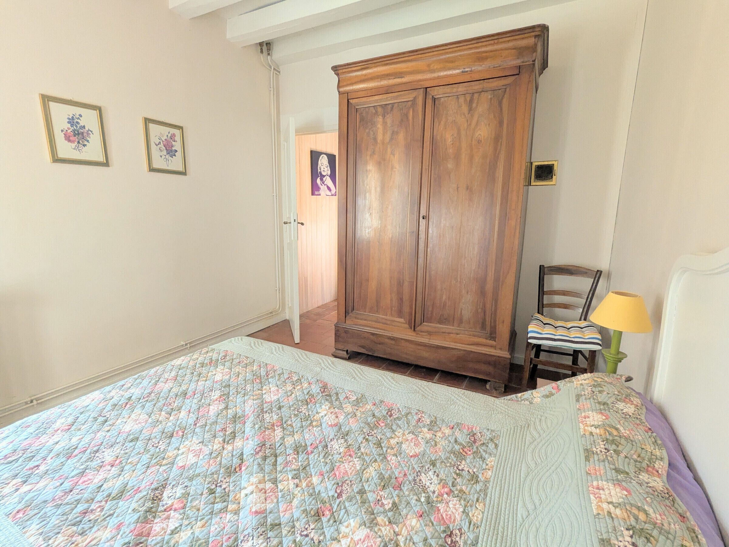 A bedroom with a large wooden wardrobe, a bed with a floral quilt, a chair with a striped cushion, and a lamp.