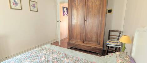 A bedroom with a large wooden wardrobe, a bed with a floral quilt, a chair with a striped cushion, and a lamp.