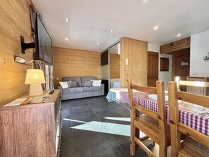 A cozy room with a wooden wall, a grey sofa, a wooden dining table with a checkered tablecloth, and a modern lamp. - Charming studio with alcove - Parking - Praz-sur-Arly (Praz-sur-Arly)