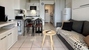 Ett kompakt bostadsutrymme med ett litet kök, en matplats med stolar, en soffa och ett litet bord med en fjärrkontroll.