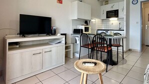 Ett kompakt bostadsutrymme med en tv på ett tv-bord, ett litet runt bord med en fjärrkontroll, ett litet kök med mikrovågsugn och spis samt en matplats med stolar.