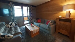 Un soggiorno accogliente con divano, tavolino e lampada. Si vede una finestra con tende e un televisore fissato al muro.
