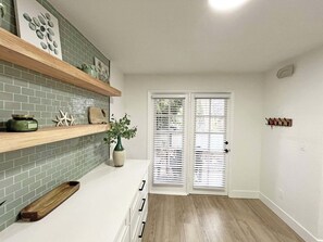 A kitchen with a green tiled backsplash, wooden shelves, a plant, and a white door with blinds. - Anastasia Oaks 104B Lo Tide (St. Augustine)
