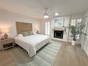 A bedroom with a bed, nightstands, a ceiling fan, a fireplace, and a large window. - Anastasia Oaks 104B Lo Tide (St. Augustine)