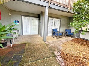 A covered patio area with two rocking chairs, a potted plant, and a brick pathway leading to a door. - Anastasia Oaks 104B Lo Tide (St. Augustine)