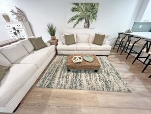 A living room with a sectional sofa, a coffee table with decorative items, a patterned rug, and a wall-mounted painting. - Anastasia Oaks 104B Lo Tide (St. Augustine)