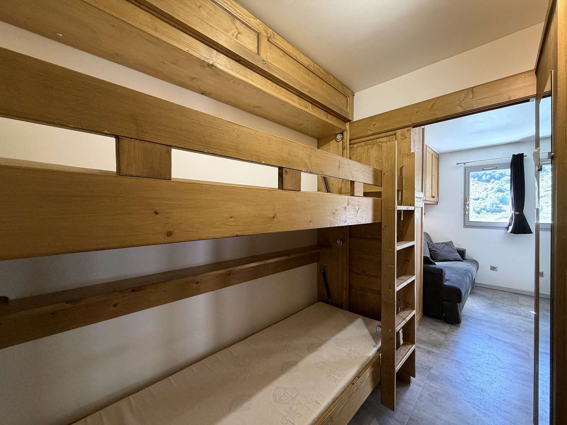 Un lit superposé avec une structure en bois et un matelas, un canapé gris et une fenêtre donnant sur des arbres.