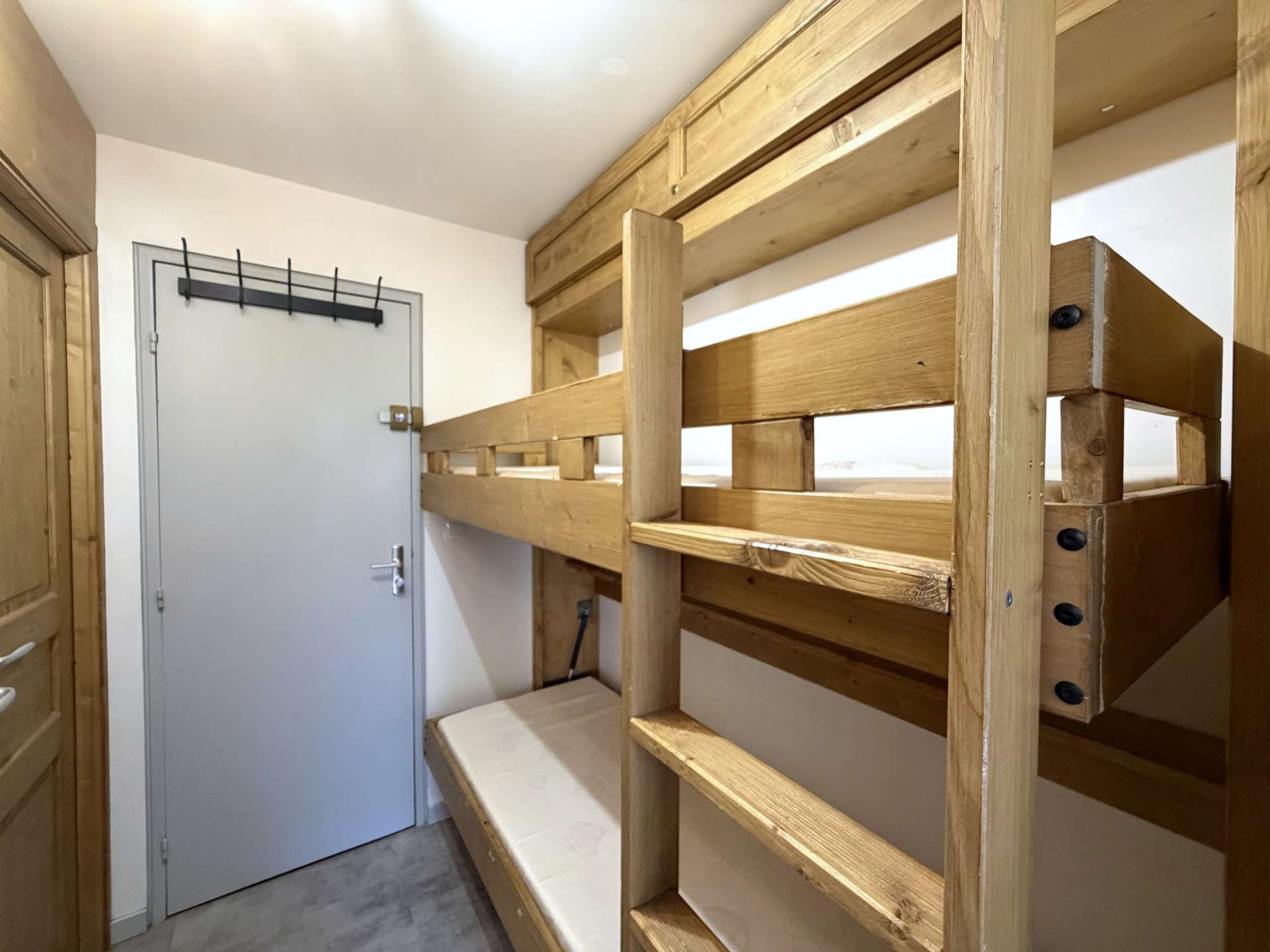 Un lit superposé avec une structure en bois et un matelas, une porte avec une poignée, et une étagère fixée au mur, sur laquelle sont posés divers objets.