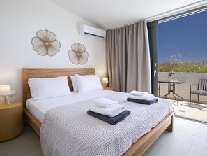 A hotel room with a large bed, a bedside table with a lamp, a small table with a chair, and a balcony with a view. - Amber by the sea – Elegant Villa with amazing view and XLarge Infinity Pool (Afteli)