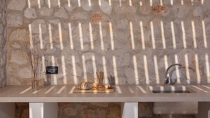 Paredes de pedra com uma bancada de cozinha, pia e tábua de corte com pão e utensílios.