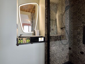 A bathroom with a mirror, sink, and shower. There is a shelf with toiletries.