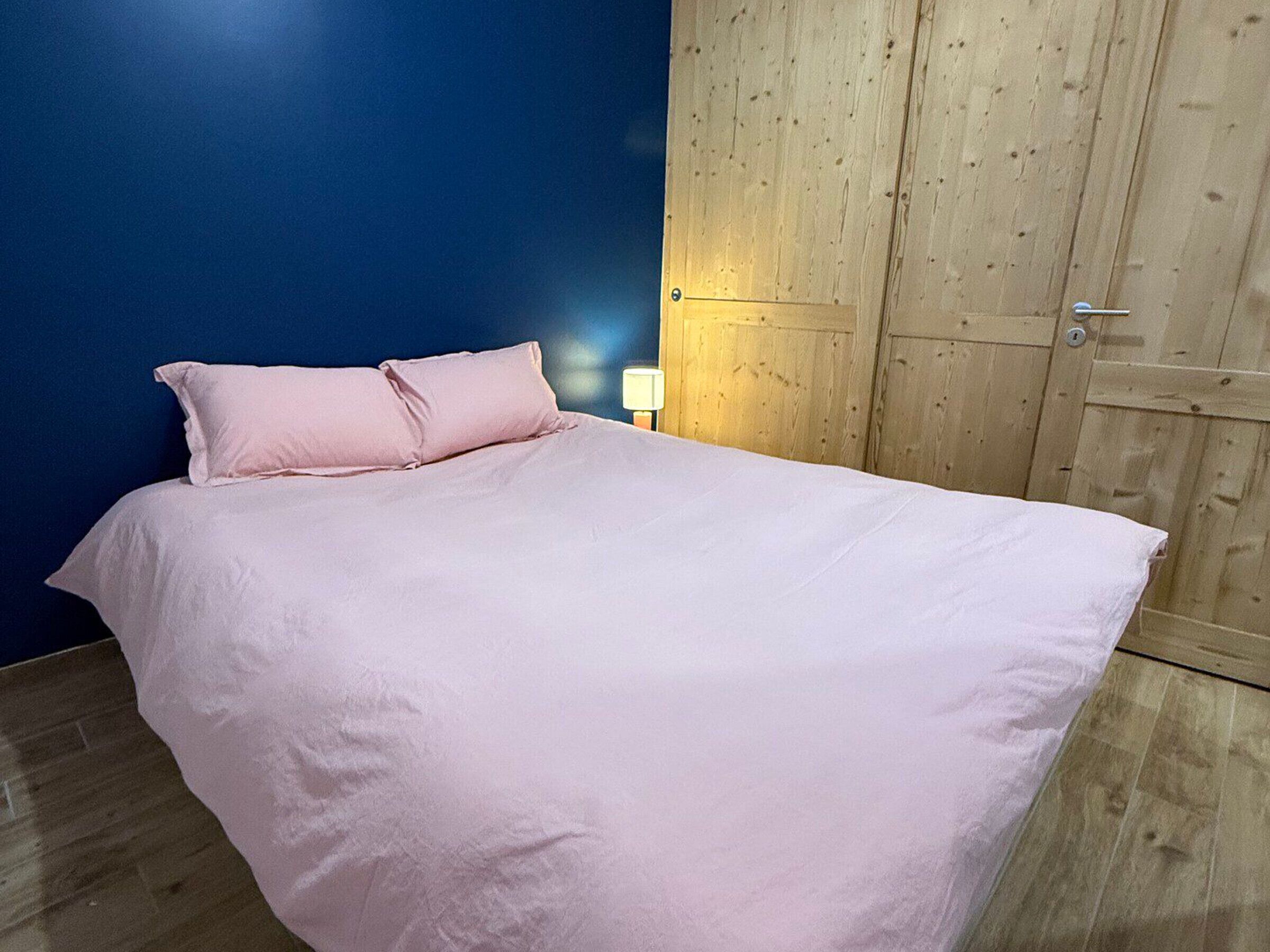 A single bed with a white mattress and pink pillows in a room with a blue wall and wooden wardrobe.