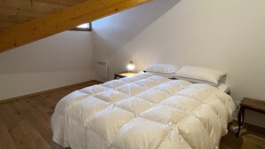 Un dormitorio con techo inclinado, cama con ropa de cama blanca, mesita de noche con lámpara y una ventana.