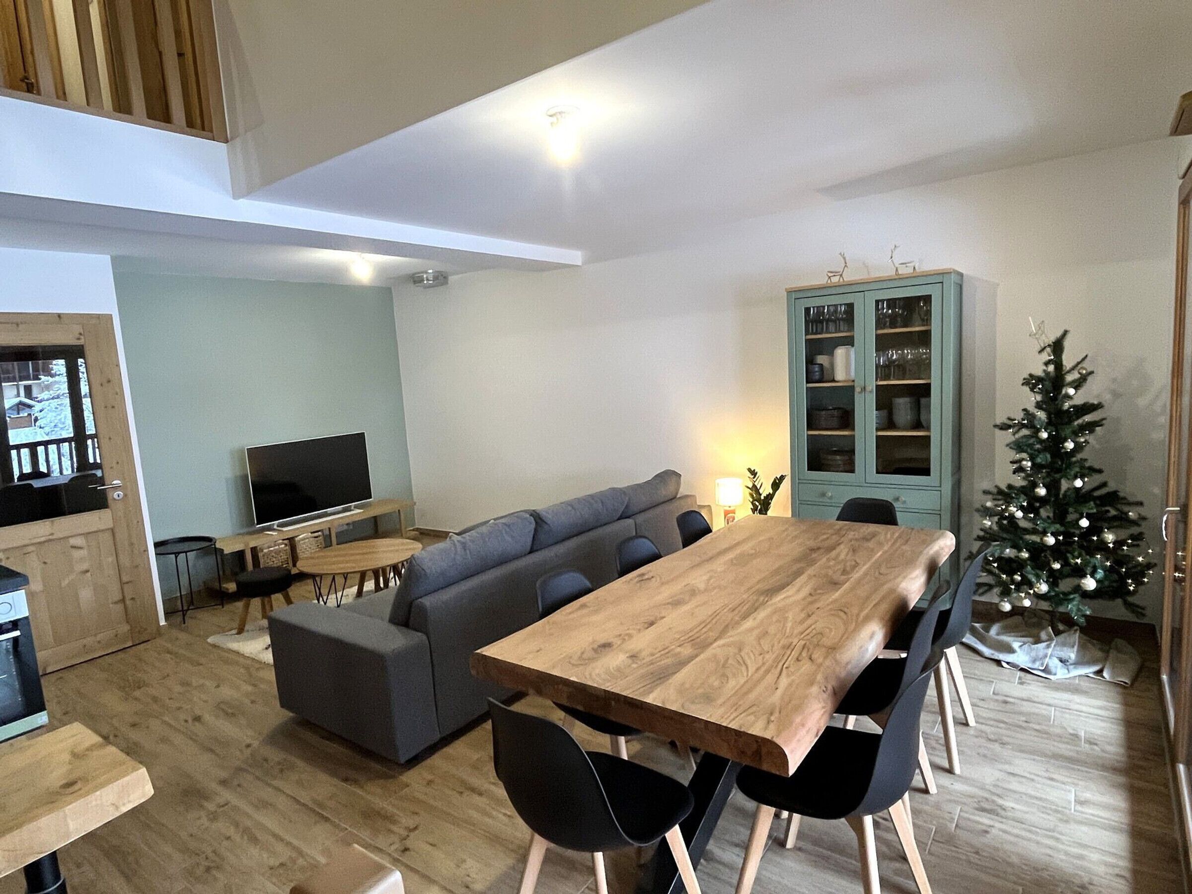 A modern living room with a wooden dining table, grey sofa, and a small TV.